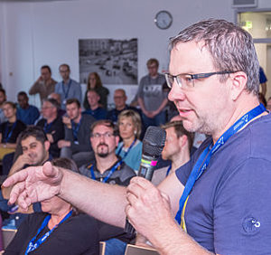 Ein Mann im blauen Hemd mit Mikro in der Hand, spricht. Im Hintergrund viele Menschen auf Stühlen, Versammlung.