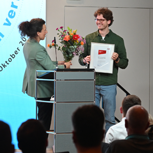 Die Selbsthilfe junger Stotternder wurde mit dem Hertie-Preis geehrt. Szene einer Preisverleihung. Eine Frau im Blazer überreicht Blumen und Urkunde an einen jungen Mann mit Locken.